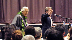 man wearing lei and another man at a microphone
